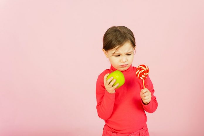 Crianças de blusa vermelha segura um pirulito em umamão e uma maçã na outra, em referência à diabetes infantil
