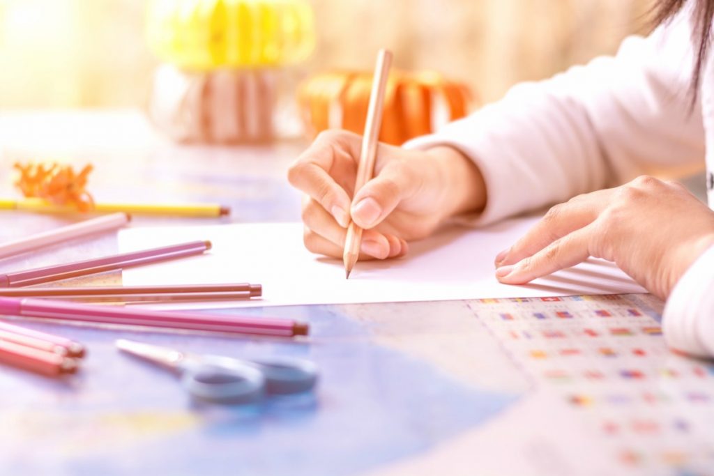 Braço de criança escreve sobre papel, rodeado por lápis coloridos e tesoura. Os desenhos e a escrita são uma atividade importante para o desenvolvimento integral das crianças.