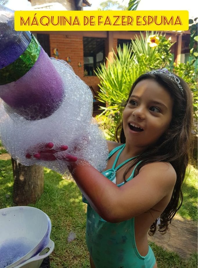 Garota de maiô está no jardim e olha feliz para brinquedo reciclado com garrafa pet que faz espuma com água e sabão.