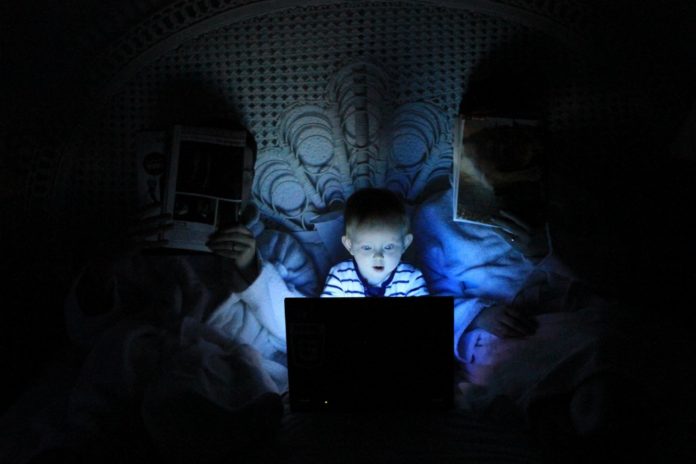 Imagem está escura e mostra pais lendo em uma cama de casal, com uma criança novinha entre eles olhando para um notebook. O tempo de tela das crianças na quarentena pode preocupar os pais.