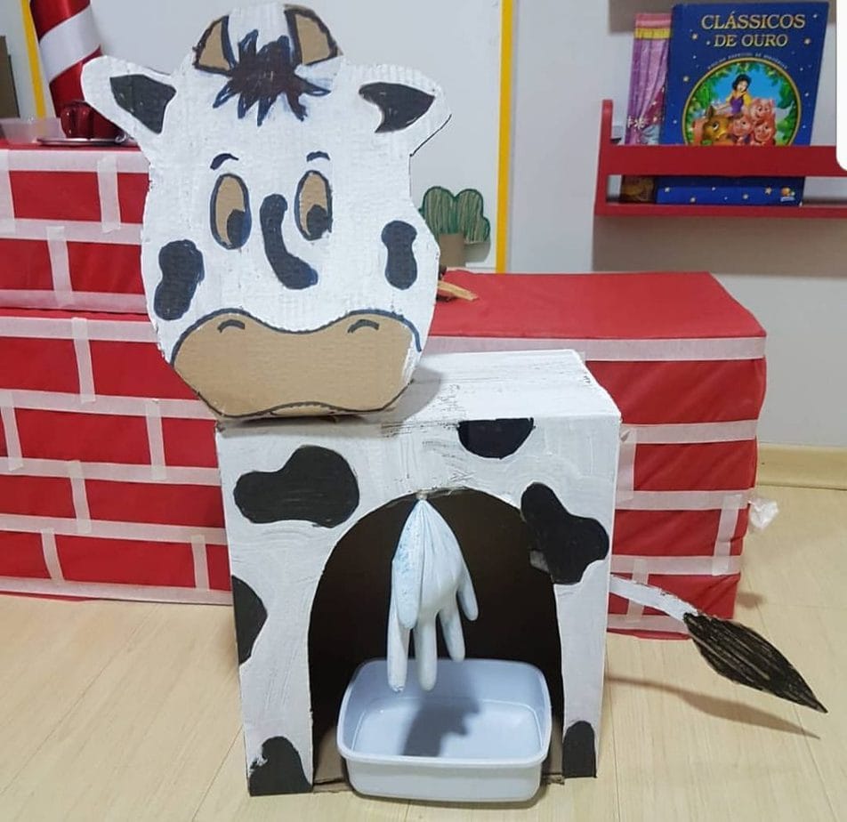 Caixa de papelão se transforma em vaquinha banca com manchas pretas e  uma luva descartável remete às tetas da vaca para que a criança brinque de tirar leite dela.