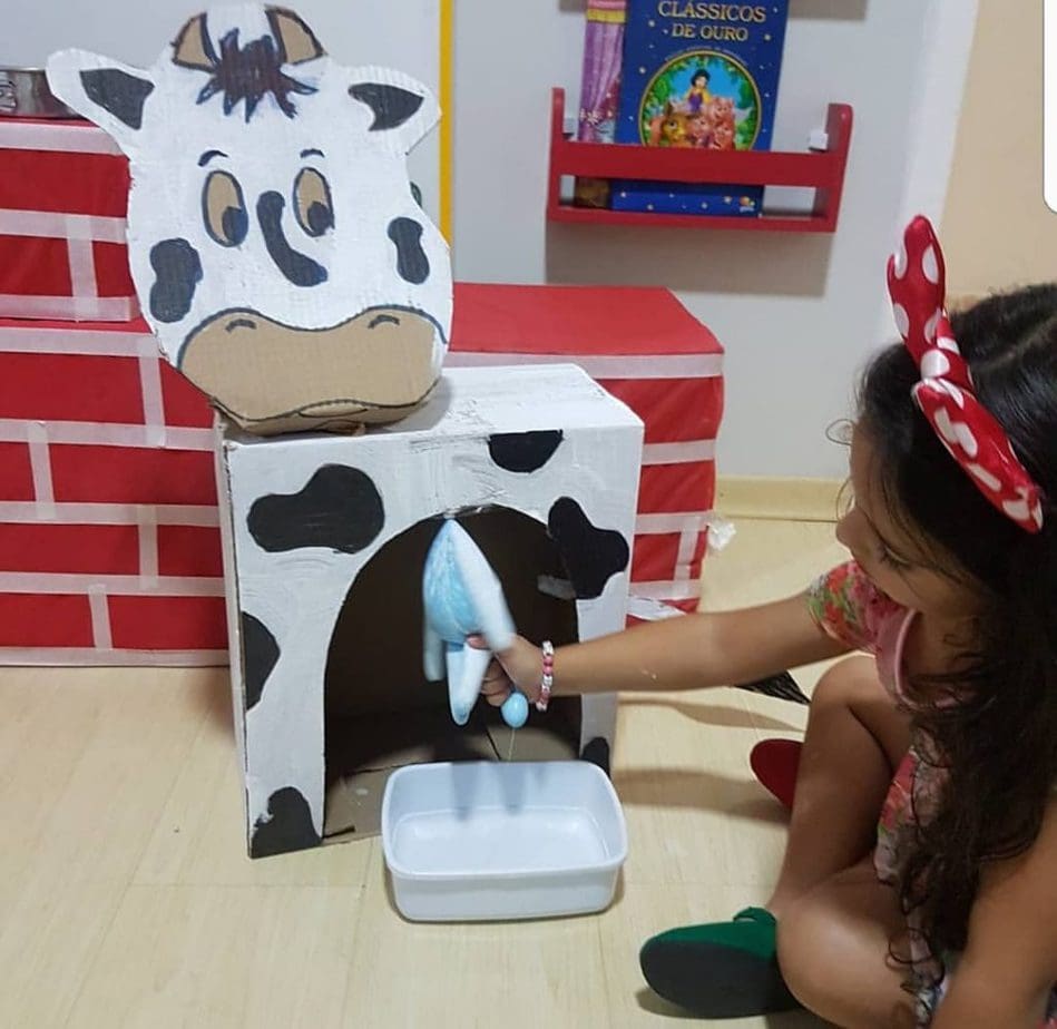 Caixa de papelão se transforma em vaquinha banca com manchas pretas e uma luva descartável remete às tetas da vaca. Criança aparece na imagem retirando "leite" da vaquinha de brinquedo.