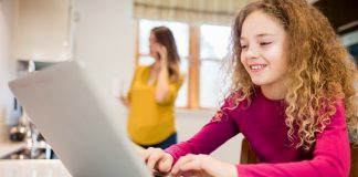 Criança usa o computador enquanto mãe está ao fundo; imagem ilustra matéria sobre acesso de crianças e adolescentes à internet.