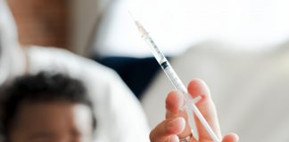 Mão segura seringa; ao fundo, bebê aguarda - imagem ilustra matéria sobre prorrogação da campanha de vacinação contra gripe.