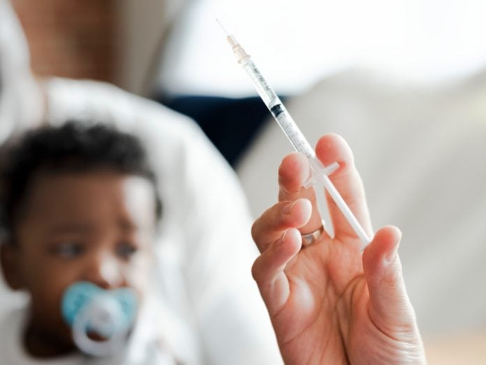 Mão segura seringa; ao fundo, bebê aguarda - imagem ilustra matéria sobre prorrogação da campanha de vacinação contra gripe.