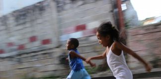 Escolas de concreto, que poderiam ser como essa imagem – em que duas crianças negras correm em um piso de cimento com uma parede ao fundo acimentada também – não incentivam o brincar e o cuidar