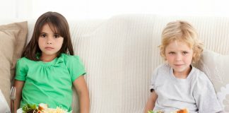 Excesso de uso de telas por crianças, como essas da imagem que comem no sofá olhando a TV, pode prejudicar capacidades motoras, sono e atividade física