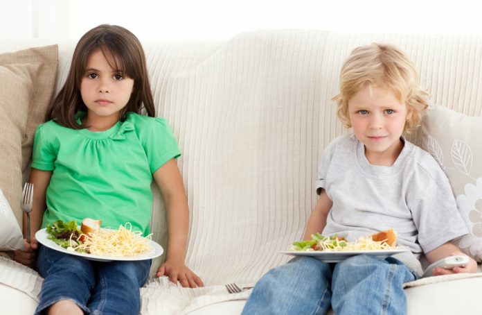 Excesso de uso de telas por crianças, como essas da imagem que comem no sofá olhando a TV, pode prejudicar capacidades motoras, sono e atividade física