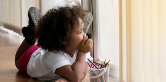 Imagem de menina desenhando e olhando pela janela pensativa, em ilustração à matéria sobre mudanças de comportamento das crianças durante a pandemia.