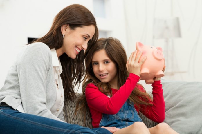 Dar mesada à criança, como garota da imagem que segura cofrinho junto à mãe, a ensina a lidar com conceitos importantes da sua vida financeira, entre os quais, a definição de orçamentos para os gastos.