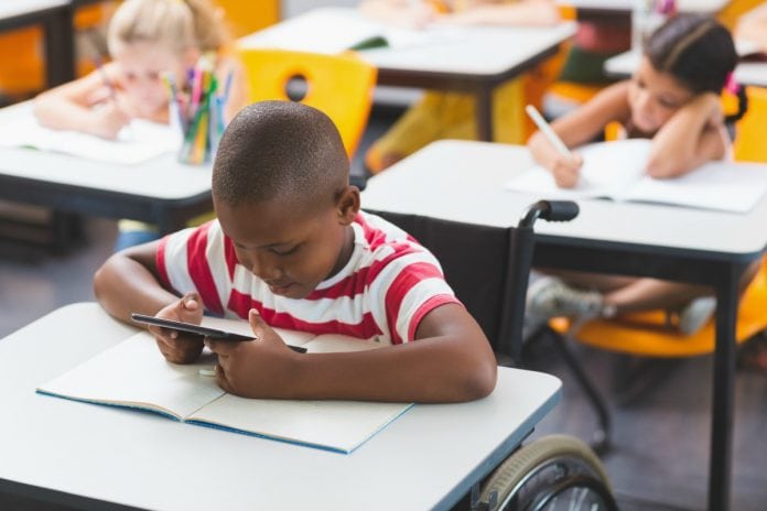 Estudantes com deficiência sem comorbidades podem voltar às aulas quando as escolas reabrirem; na imagem garoto negro e cadeirante está em sala de aula junto com outros alunos olhando tablet sobre a mesa