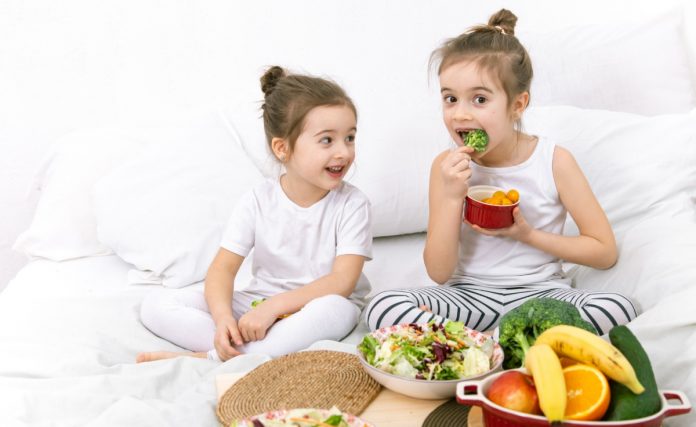 Duas crianças em meio a travesseiros e pratos de salada e frutas; imagem ilustra matéria com dicas sobre como manter a alimentação das crianças saudável.