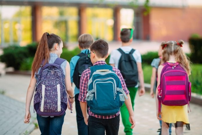 Crianças de costas, usando mochilas, andam em direção à escola; imagem ilustra matéria sobre coronavírus nos Estados Unidos.