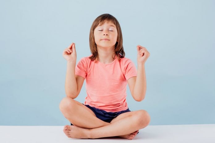 Meditação mindfulness: 3 dicas para começar a práticar com as crianças; na imagem menina respira sentada com pernas cruzadas, olhos fechado e antebraços suspensos