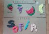 Brincadeiras com massinha: desenhando frutas na caixa de papelão; imagem mostra papelão com frutas e legumes desenhados e a massinha colorida por cima deles, no meio, nome Sofia escrito e coberto de massinha
