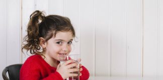 Doenças de inverno: 6 cuidados para evitar que as crianças adoeçam; na imagem, criança de blusa vermelha segura copo d'água sorridente