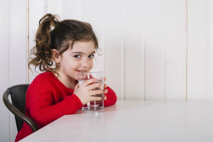 Doenças de inverno: 6 cuidados para evitar que as crianças adoeçam; na imagem, criança de blusa vermelha segura copo d'água sorridente
