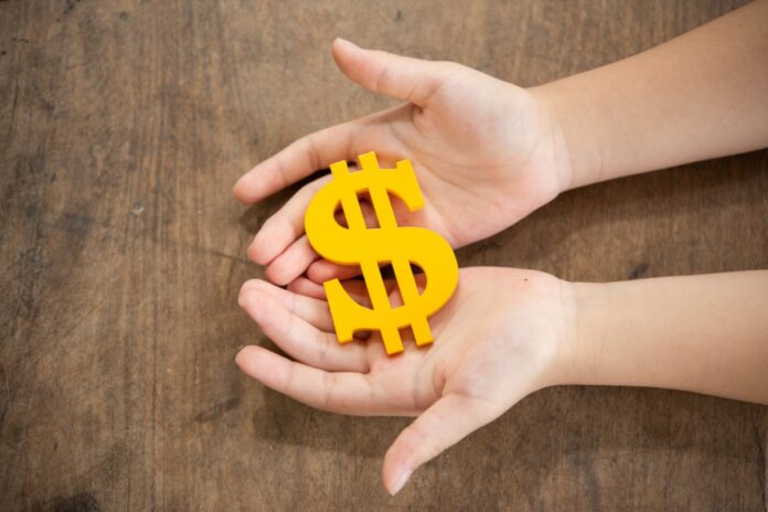 Duas mãos seguram um cifrão amarelo em ilustração à matéria sobre crianças com conta em banco.