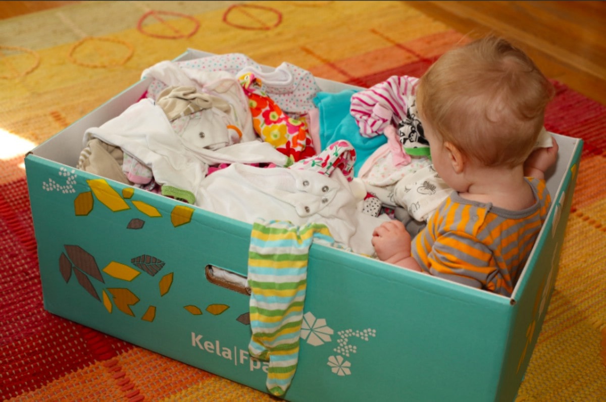 Bebê sentado dentro de uma caixa azul com roupas infantis a sua volta.