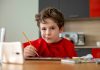 Aulas online e maternidade atípica; na imagem, menino de blusa vermelha pinta algo em frente a um tablet e faz bico com a boca para o lado