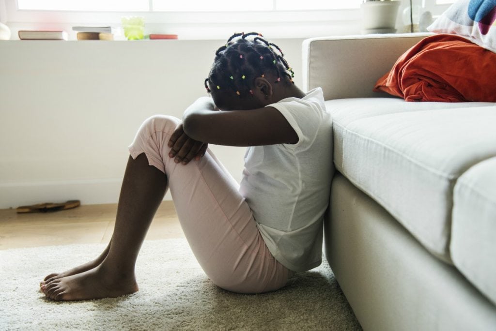 Depressão nas crianças: como identificar para evitar chegar ao suicídio; na imagem, garota negra está sentada no chão com costas no sofá e cabeça sob os joelhos dobrados