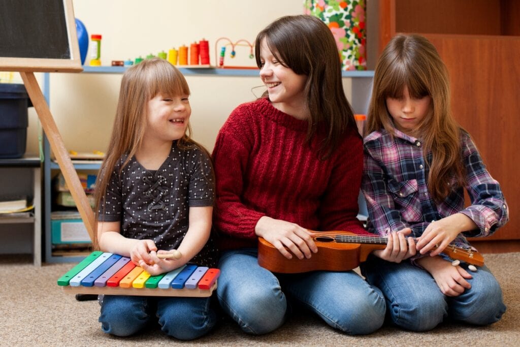 10 sugestões para falar sobre deficiência e inclusão com as crianças; imagem mostra três meninas sentadas brincando com instrumentos musicais Uma delas tem síndrome de Down e tem cabelos loiros e pele clara. As outras duas têm cabelos castanhos, também com a pele clara.