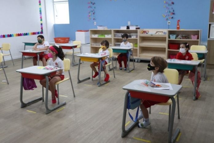 Em sala de aula, crianças, de máscara, sentam em carteiras distantes umas das outras; imagem ilustra matéria sobre aulas presenciais em escolas particulares.