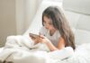 Compras online: crianças gastam mais do que adolescentes, revela estudo; imagem mostra garota de cabelo comprido mexendo no celular sentada na cama de lençóis brancos