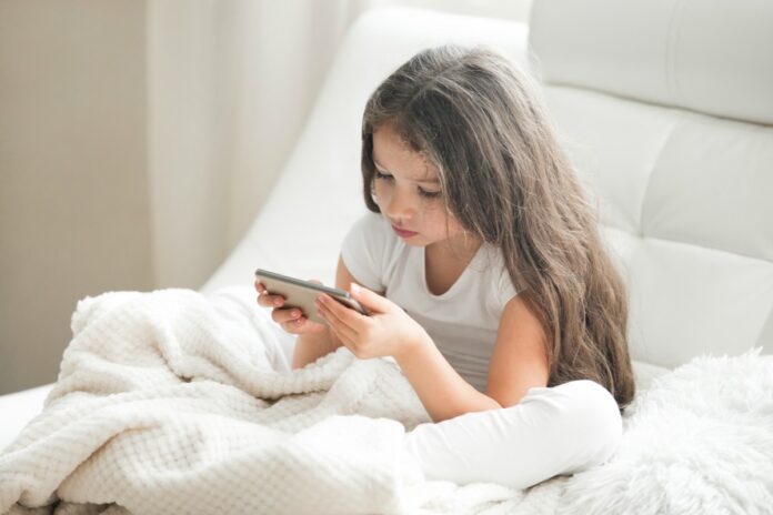 Compras online: crianças gastam mais do que adolescentes, revela estudo; imagem mostra garota de cabelo comprido mexendo no celular sentada na cama de lençóis brancos