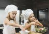 3 receitas que as crianças adoram (e usam alimentos da época); na imagem, duas meninas com chapéu de cozinheiro e avental preparam salada
