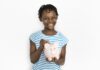 Semanada ou mesada: qual é o mais indicado para o seu filho?; imagem mostra garota de camiseta listrada azul sorridente segurando cofrinho de porquinho cor-de-rosa