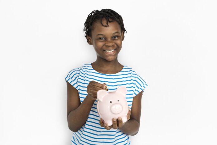 Semanada ou mesada: qual é o mais indicado para o seu filho?; imagem mostra garota de camiseta listrada azul sorridente segurando cofrinho de porquinho cor-de-rosa