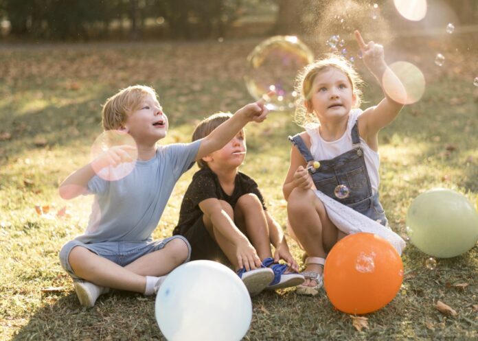 Criança deve ser, antes de tudo, criança; imagem mostra três crianças sentadas na grama brincando com bexigas e bolhas de sabão