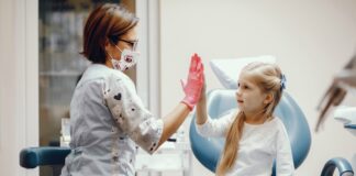 Em casa de urgência médica, escolha um atendimento humanizado; imagem mostra médica de máscara batendo a mão, palma com palma, com criança sentada em cama de hospital