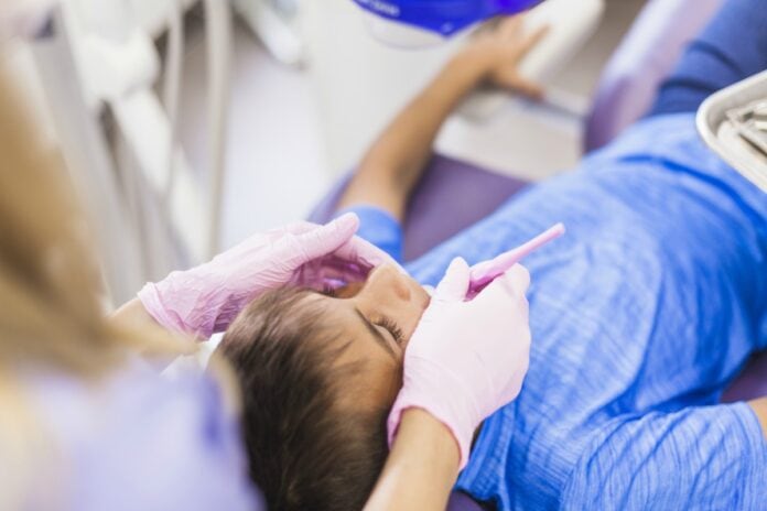 Atendimento odontológico de crianças diminui em 88% na pandemia; imagem mostra mão de dentista com luva mexendo na boca de menino sentado em cadeira de consultório