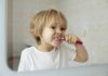 Dicas para estimular a escovação correta dos dentes nas crianças; imagem mostra menino de cabelos claros escovando os dentes em frente ao espelho