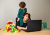 1 em cada 4 mães pensa em pedir demissão do trabalho; imagem mostra mulher sentada olhando para computador sobre a mesa e uma criança está sentada sobre seus ombros com a mão em seus cabelos