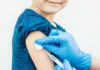 Coberturas vacinais: metade das vacinas infantis não atinge meta desde 2015; imagem mostra mão com luva azul emborrachada aplicando dose de vacina em braço de criança de blusa azul