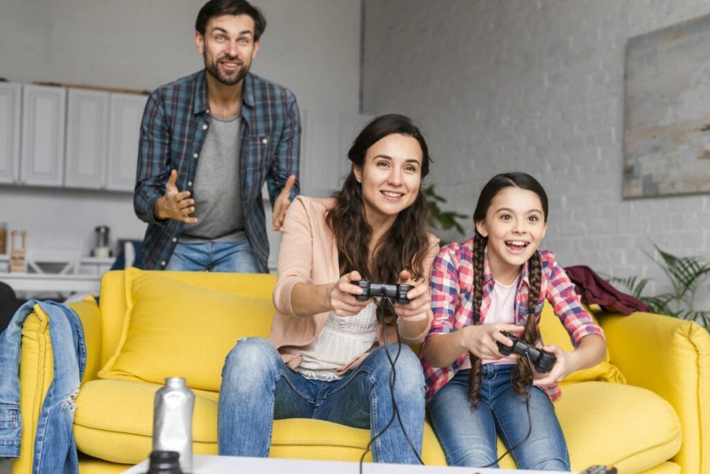 Videogames: um manual para pais e mães jogarem com os filhos (sim, isso é importante); imagem mostra mãe e filha jogando no sofá e o pai mais atrás vibrando com o jogo