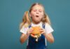 O que comemos e que comidas damos aos nossos filhos: presença de agrotóxicos em alimentos e riscos dos ultraprocessados despertam alerta; menina come hamburguer