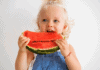 Para ajudar na digestão: 7 alimentos que evitam a constipação na criança; imagem mostra criança loira segurando e mordendo fatia de melancia