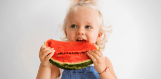 Para ajudar na digestão: 7 alimentos que evitam a constipação na criança; imagem mostra criança loira segurando e mordendo fatia de melancia
