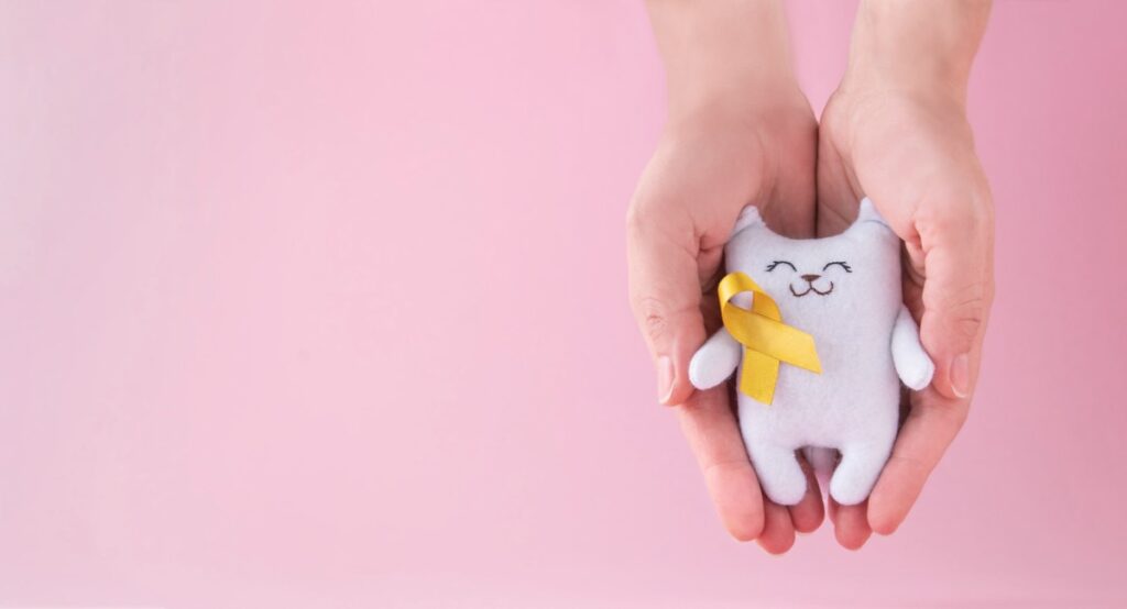 Câncer infantil: diagnóstico precoce é essencial apra aumentar as chances de cura; imagem msotra gatinho branco de tecido com laço amarelo em fundo de imagem rosa