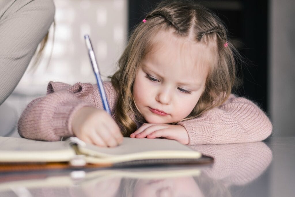 Seu filho sabe segurar o lápis corretamente? Veja como ajudá-lo; imagem mostra menina de 4 ou 5 anos escrevendo em um caderno