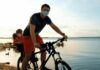 Flexibilização com segurança para as crianças: é possível?; imagem mostra pai andando de bicicleta com o filho em cadeirinha no bagageiro, ambos de máscara com lago ao fundo