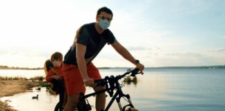 Flexibilização com segurança para as crianças: é possível?; imagem mostra pai andando de bicicleta com o filho em cadeirinha no bagageiro, ambos de máscara com lago ao fundo