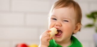 A primeira papinha: como se saber já é hora de ofertar alimentos ao bebê; bebê de babador verde segura sorridente banana na boca