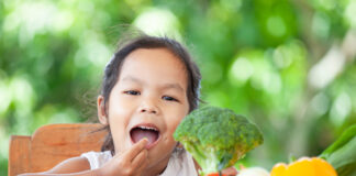 Índice de anemia cai pela metade em crianças de até cinco anos; menina morena segura brocolis em uma mão e na outra leva à boca um pedacinho desse vegetal