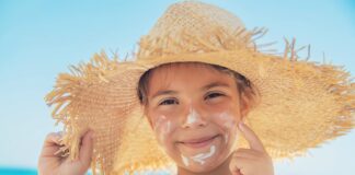 Câncer de pele: cuidados com exposição solar começam na infância; garoto passa protetor no rosto e usa chapéu de palha