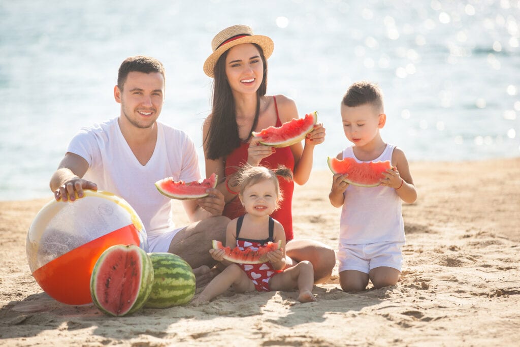 Veja opções de lanches caseiros rápidos para as crianças neste verão; família sentada na areia da praia comendo melancia